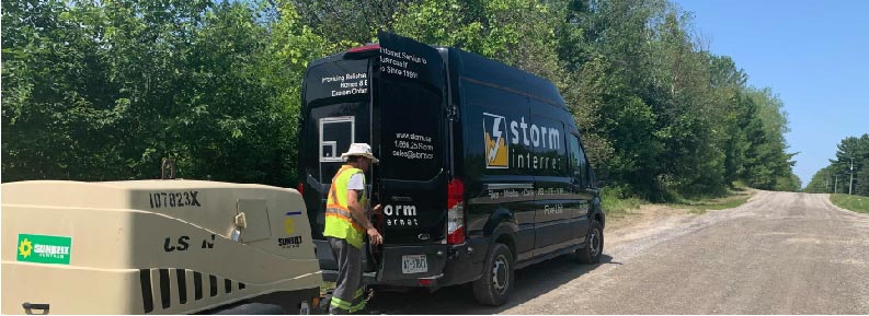 A field technicial opening the back of a Storm Internet van. There is a generator being towed by the van. The van is parked on the side of a dirt road. The technician is surrounded by trees on a clear, sunny day.