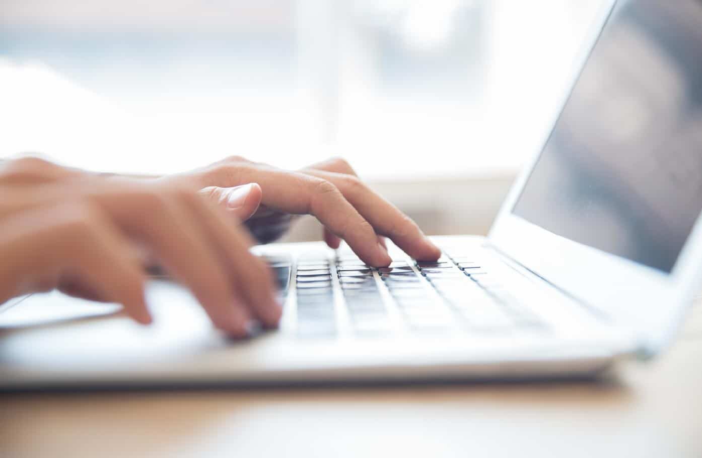 A close up image of hands typing on a laptop in a brightly lit setting.