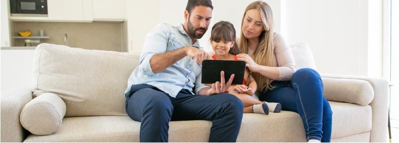 A man, woman, and their child are sitting on a couch. The man is holding a tablet for all of them to see.