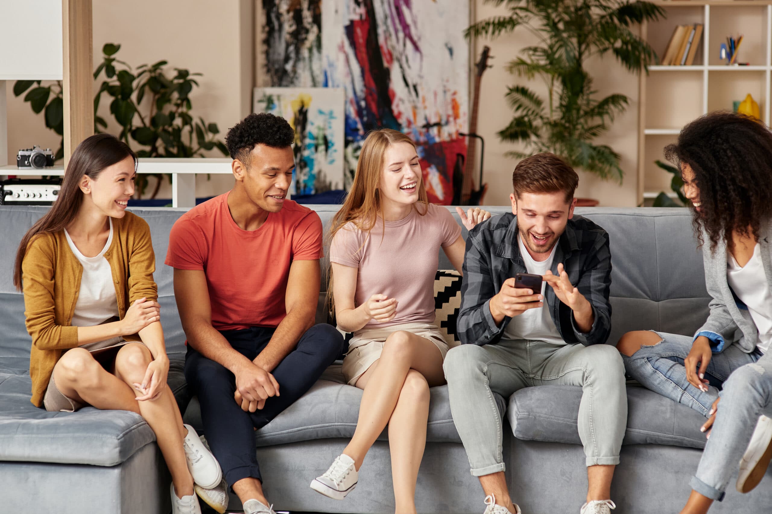Group of happy young multicultural people in casual wear looking at smartphone, enjoying time together while sitting on the sofa in the living room. Leisure concept