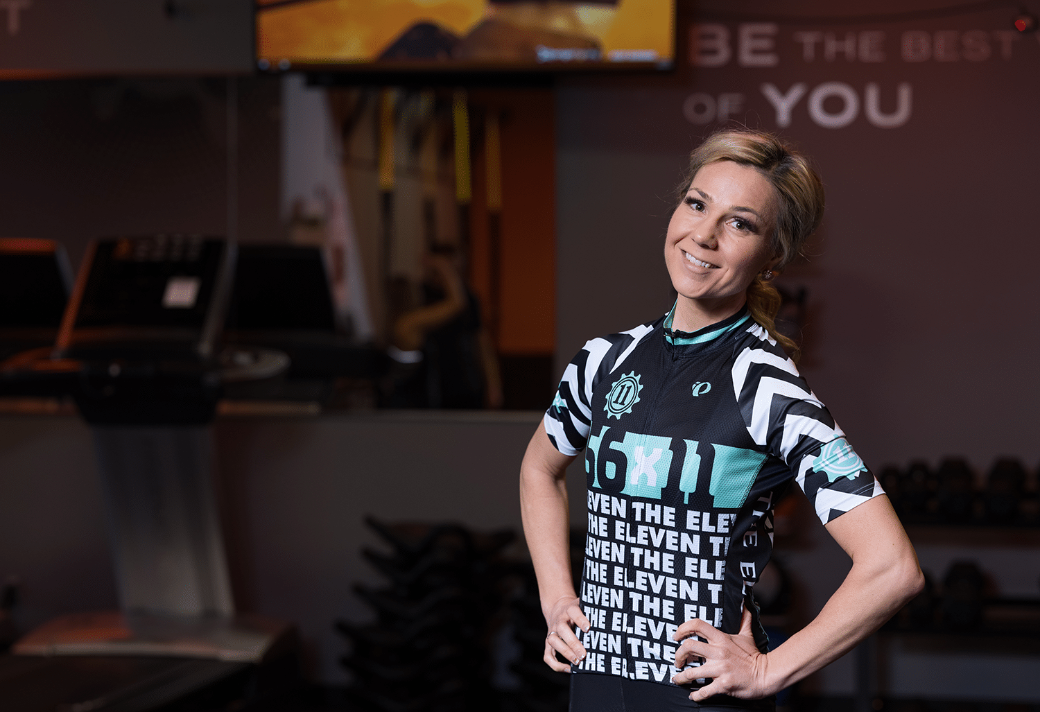 Angella Gorad posing for the camera in a cycling outfit in a gym setting. A treadmill and some free weights are displayed in the background.